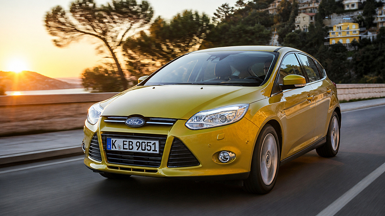 Yellow Ford Focus hatchback driving on a road at sunset with trees and buildings in the background.