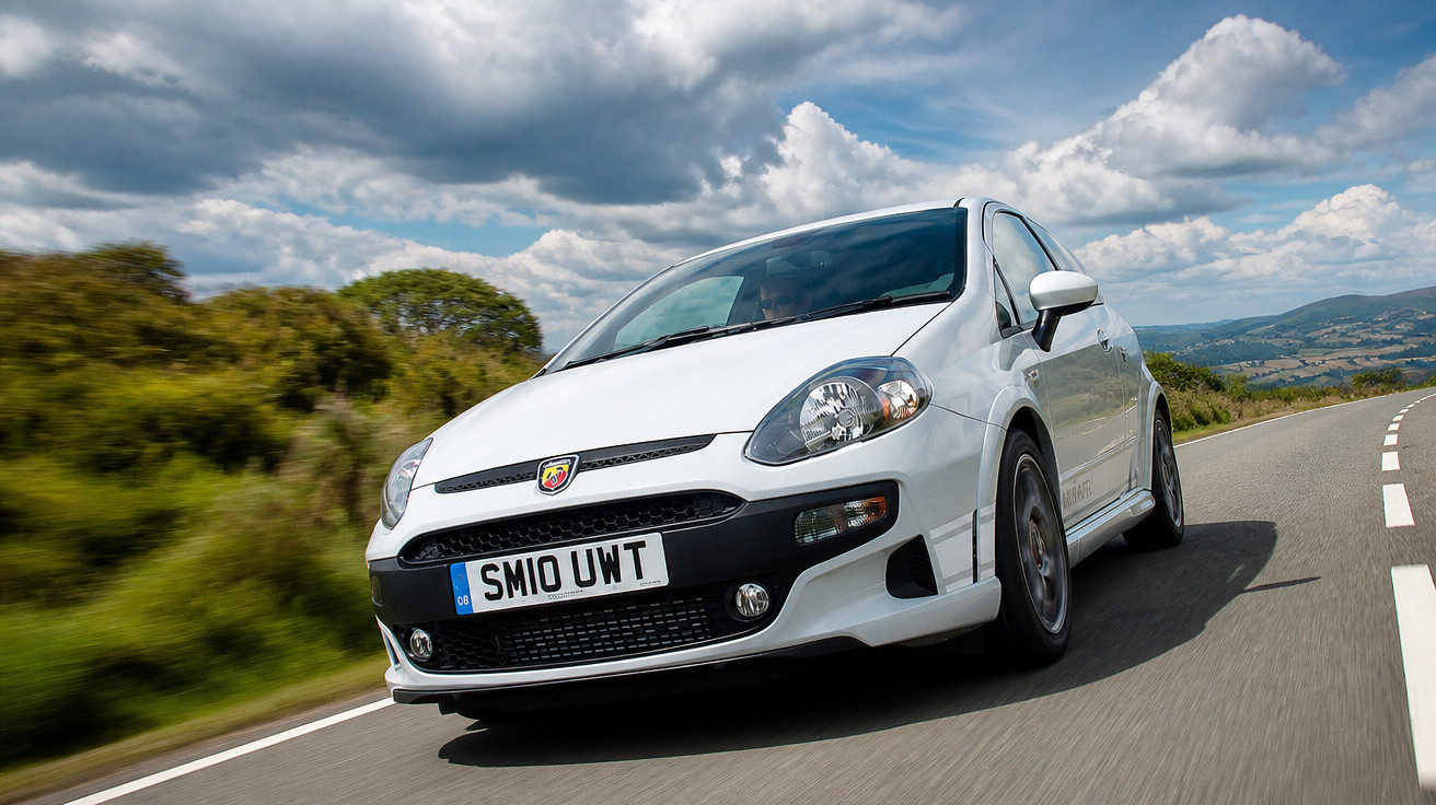 White Abarth sports car driving on a winding country road under a cloudy sky.