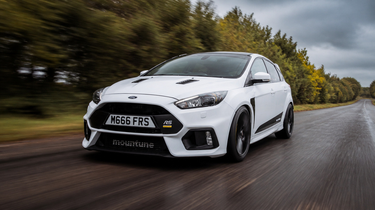 White Ford Focus RS with black wheels driving on wet country road with greenery and cloudy sky.