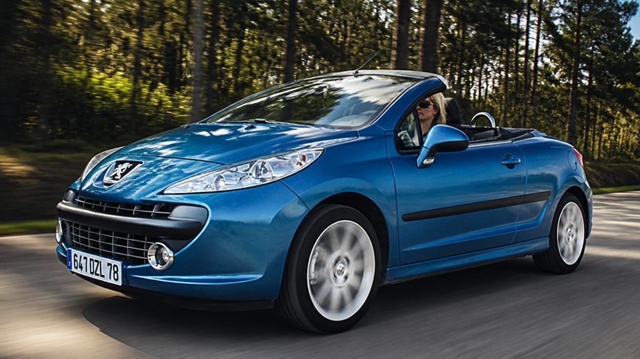 Blue Peugeot convertible driving on a forest-lined road with the top down and a driver inside.