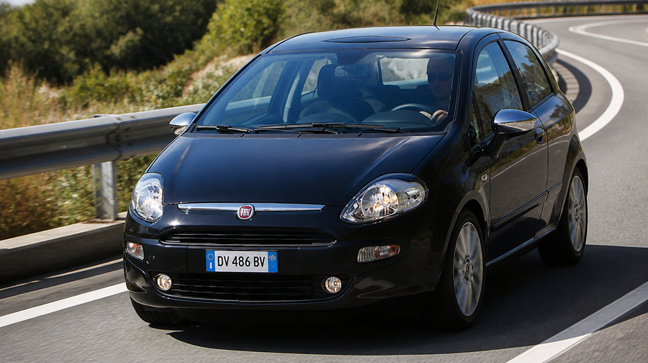 Black Fiat hatchback car driving on a curved road with metal guardrails and greenery in the background.
