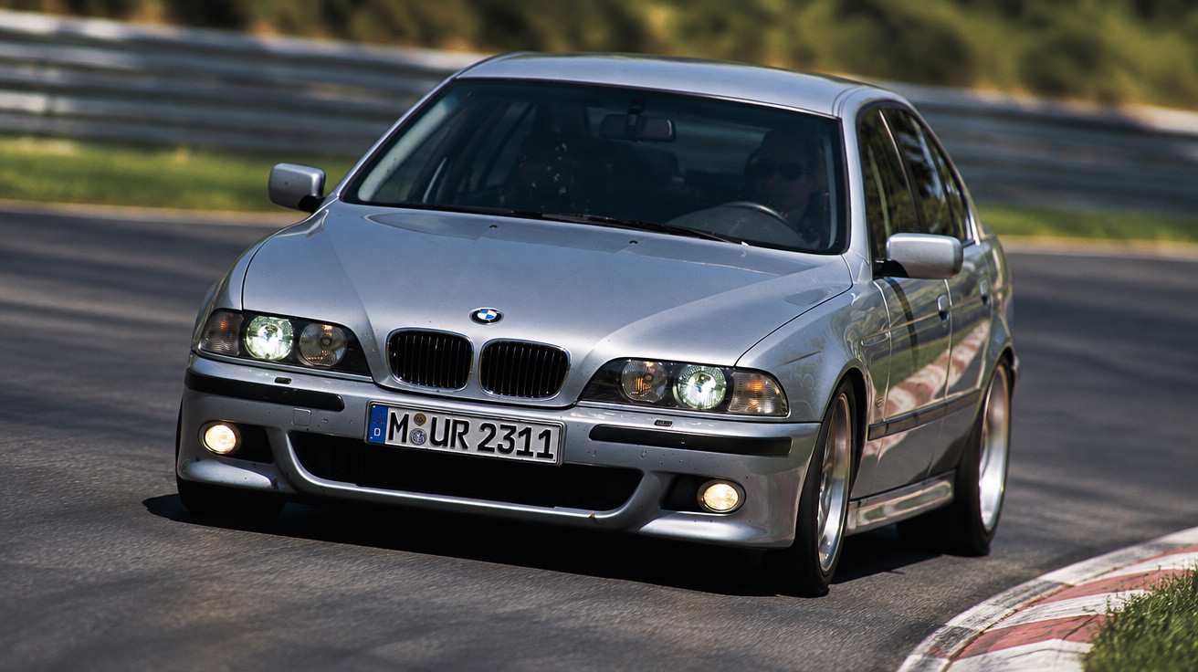 Silver BMW sedan with headlights on driving on a curved racetrack surrounded by greenery.