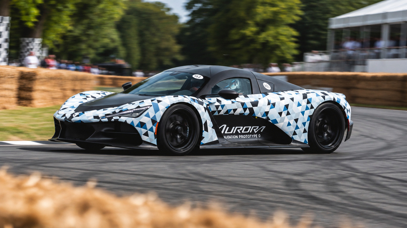 Aurora Validation Prototype 0 sports car with geometric camouflage driving on a race track with hay bales.