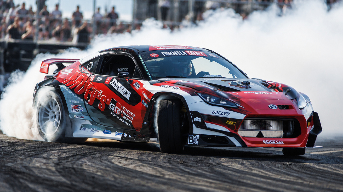 Red and black Toyota drift car sliding on a track with smoke billowing from its rear tyres during a race event.
