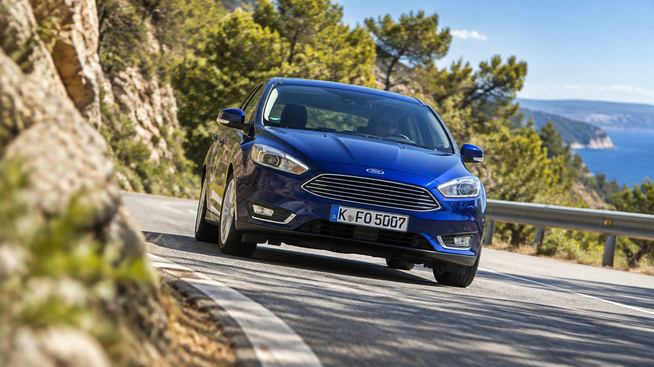 Blue Ford Focus driving on a winding coastal road with rocky cliffs and trees on a sunny day.